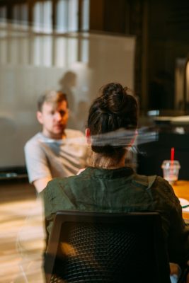Two people having a discussion in a modern office setting, with one person facing the camera and another seated across the table.